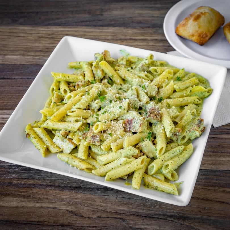 Penne pasta with pesto sauce, chicken, and parmesan cheese on a white plate with bread.