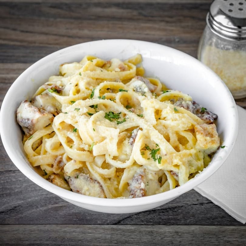 Creamy fettuccine Alfredo with chicken and parsley in a white bowl, with a cheese shaker nearby.